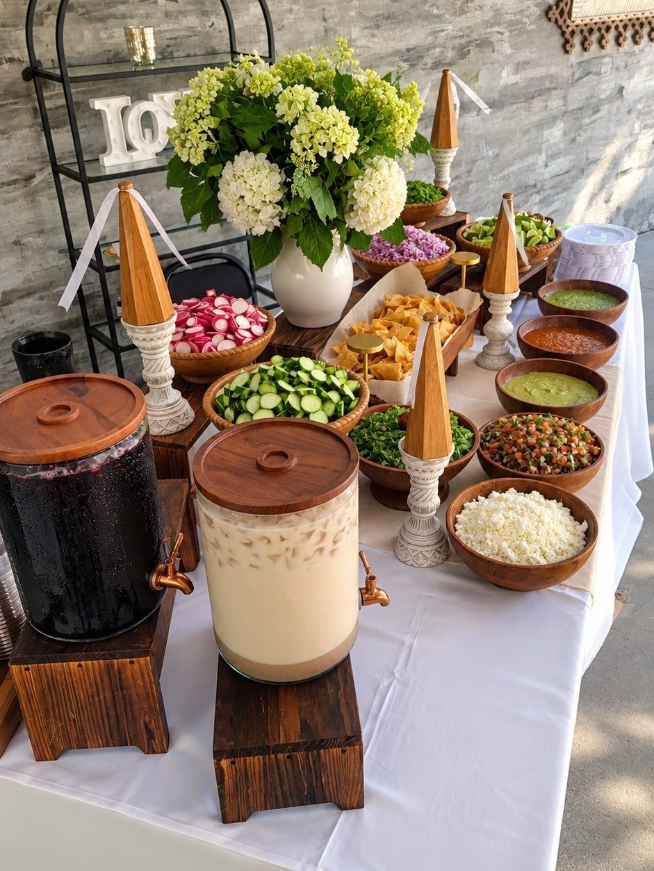 Taco bar spread with various toppings in wooden bowls and beverage dispensers.