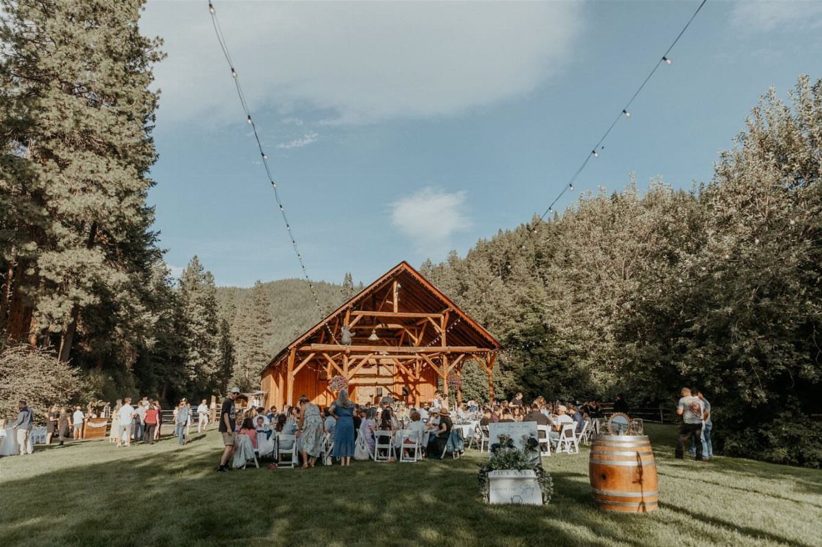 Outdoor wedding reception with guests and a wooden barn in a forested mountain setting