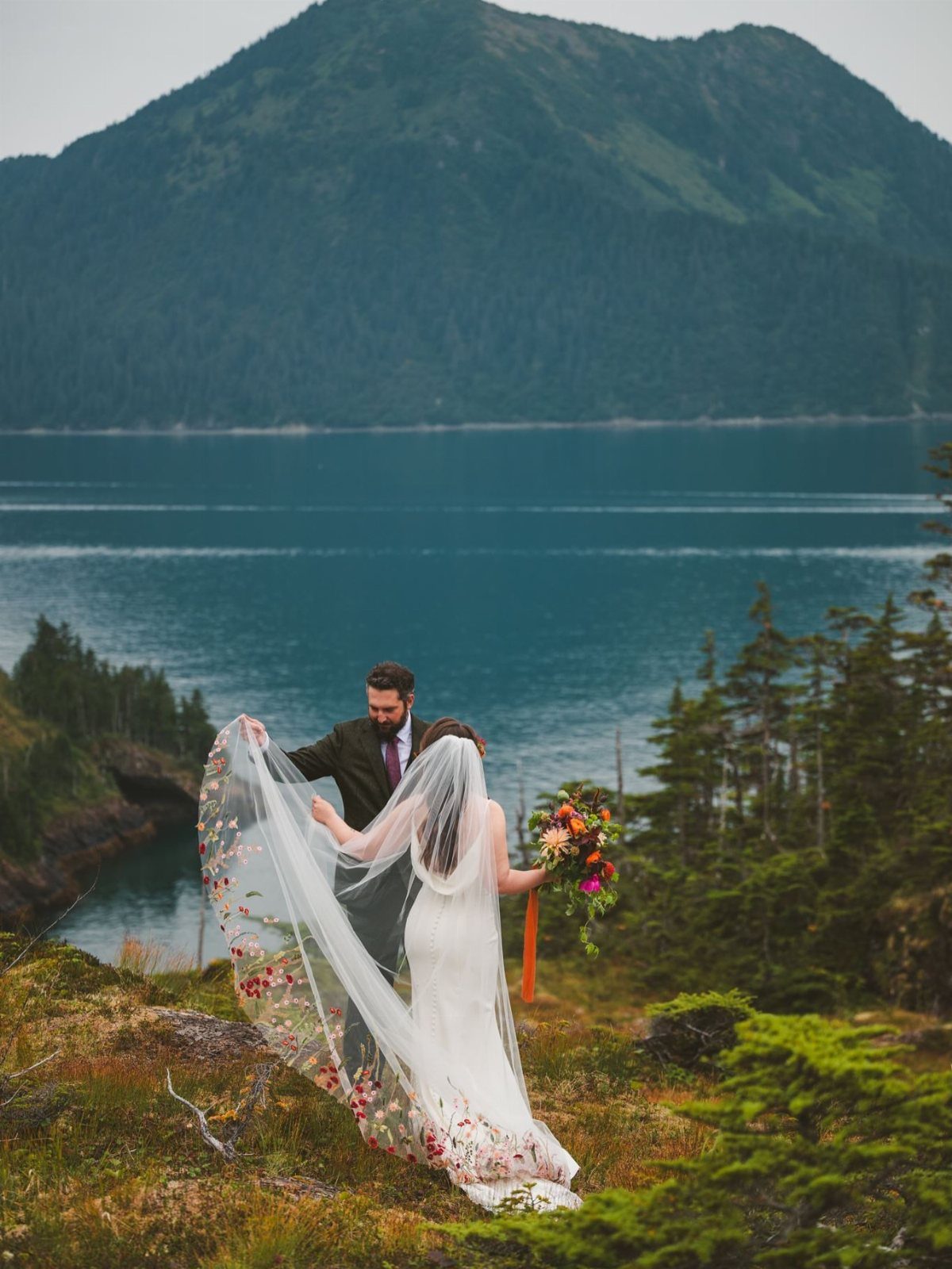 Bride and groom standing on a grassy cliff overlooking a lake and mountain.