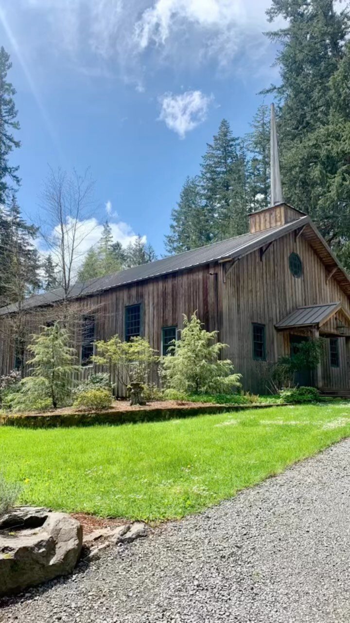Rustic wooden chapel with a steeple nestled among tall evergreen forest trees.