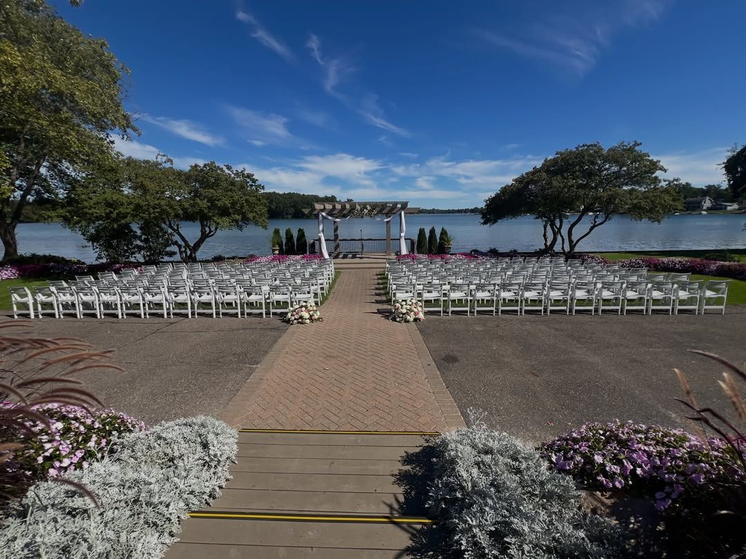 White wedding chairs and pergola overlooking a calm lake with green trees.
