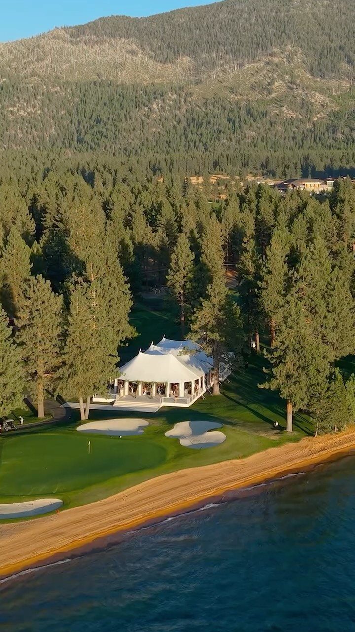 White pavilion on a green lawn by a lake and sandy beach with mountains.
