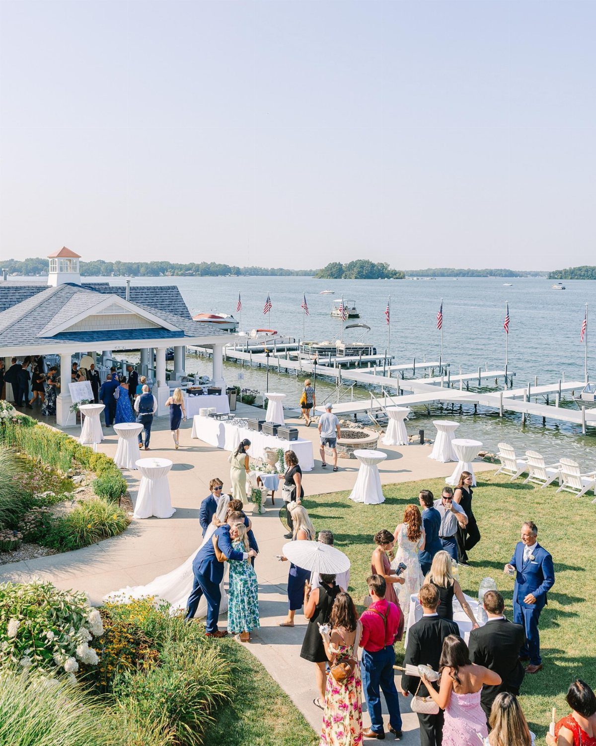 Lakeside wedding venue with guests gathering near a white pavilion and long dock.