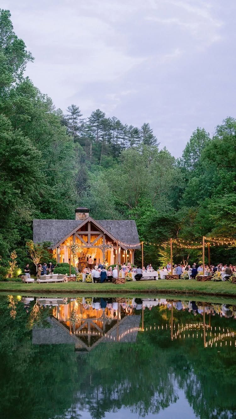 Rustic wooden lodge with string lights and guests by a reflective pond at twilight.