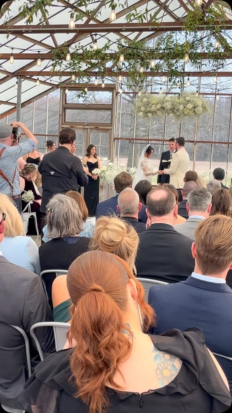 Wedding ceremony inside a glass greenhouse with guests and hanging string lights.