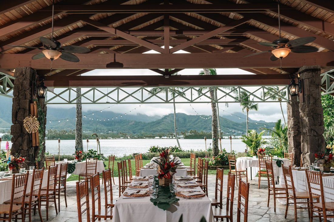 Outdoor wedding reception setup in a thatched pavilion overlooking tropical mountains and water.