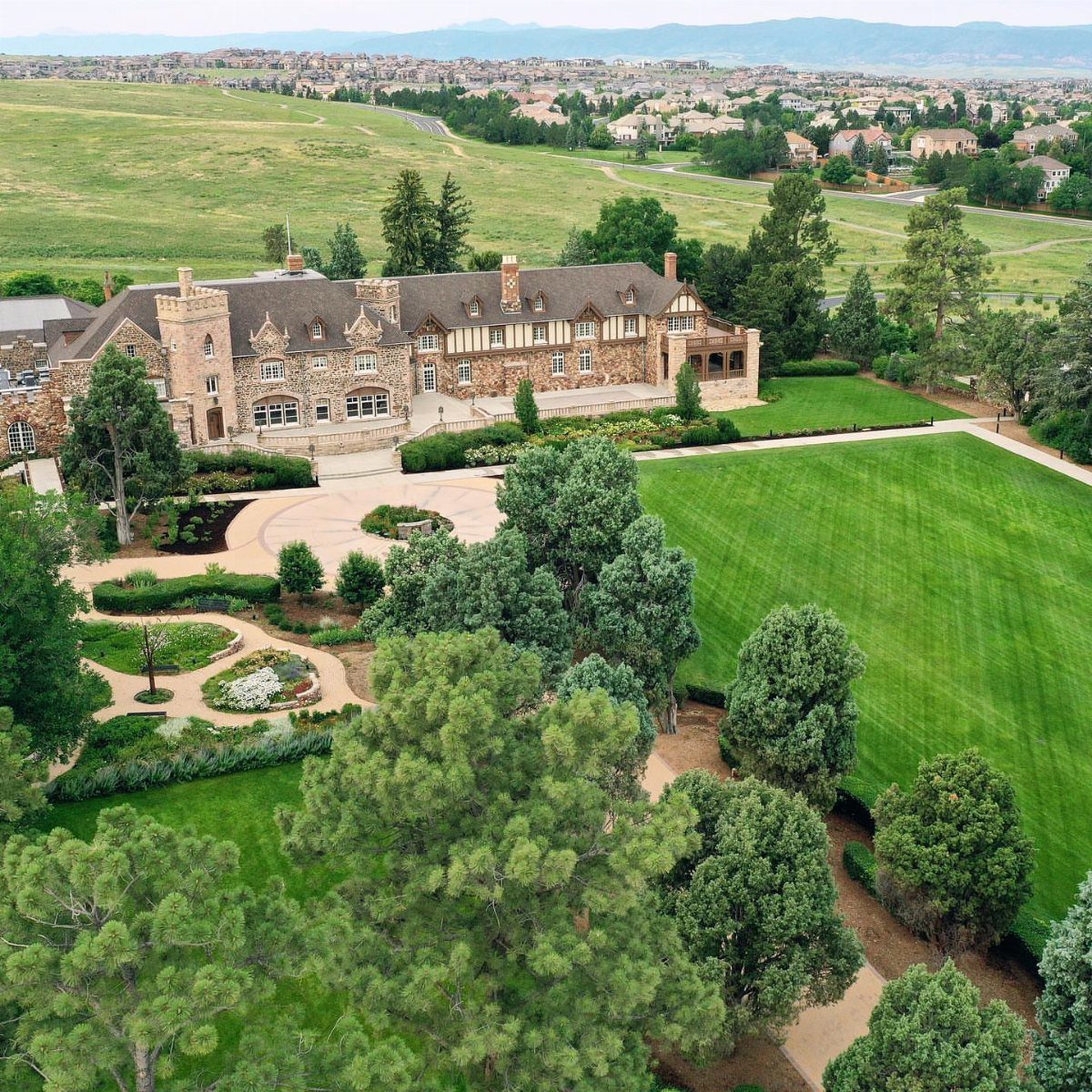 Historic stone mansion with sprawling green lawns and manicured gardens under a clear sky.