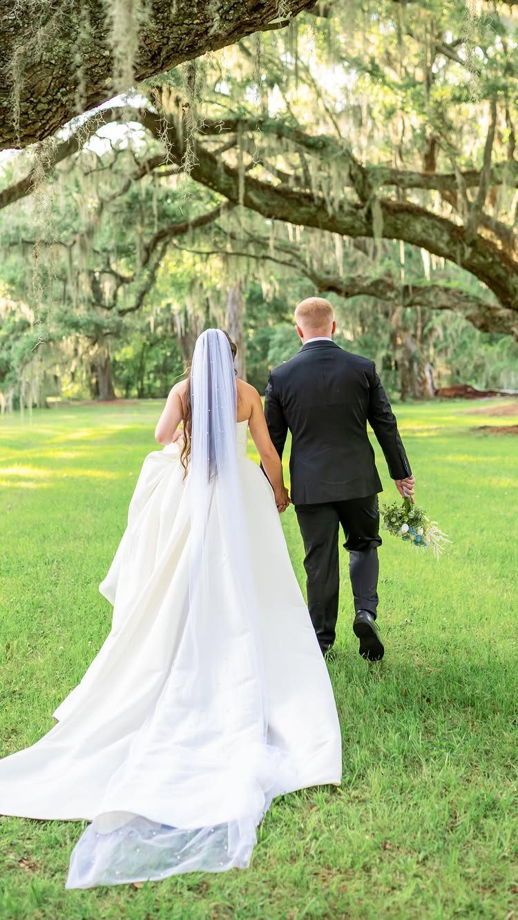 Bride and groom walking through a field under oak trees with Spanish moss.
