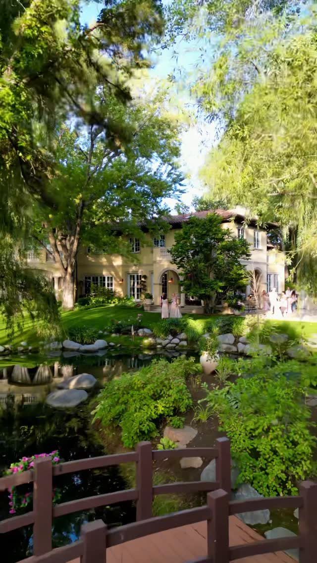 Mediterranean villa with European gardens, a pond, and a wooden bridge foreground.