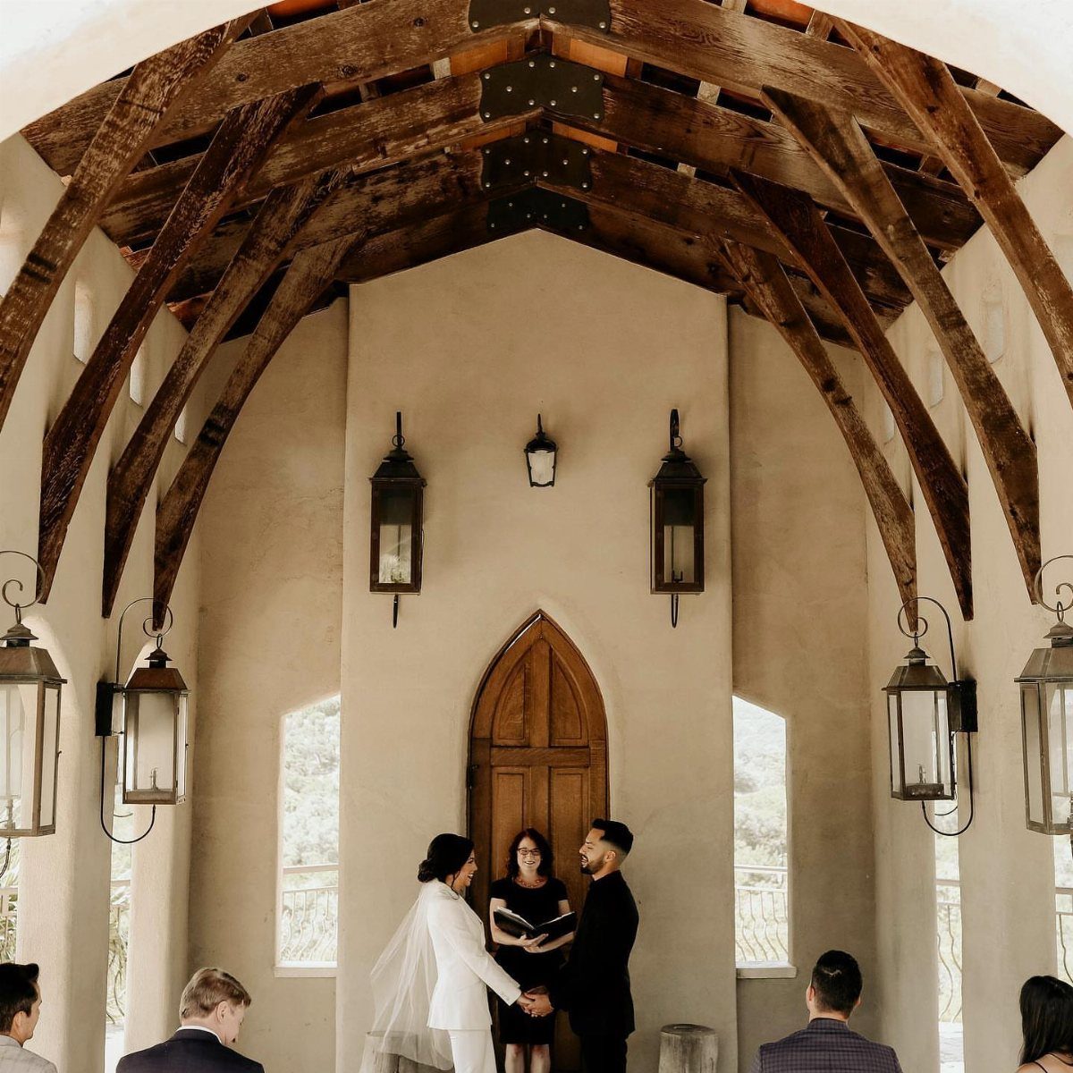 Couple holding hands during a wedding ceremony under a vaulted wooden beam ceiling.