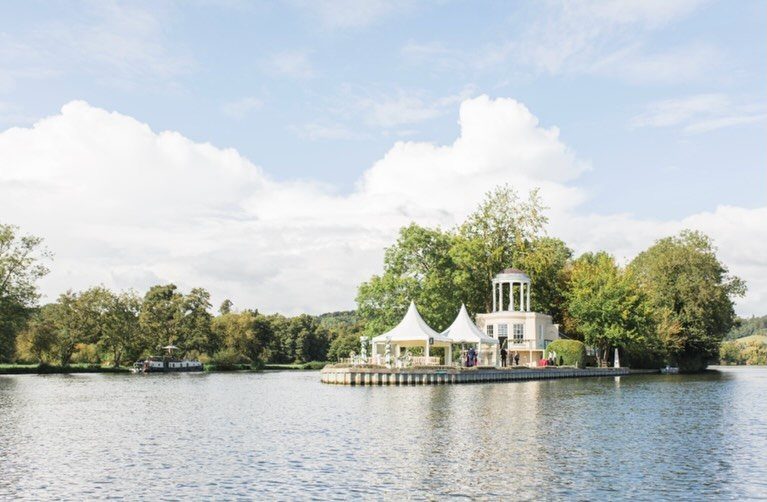 White pavilion with pointed tents and a tower on a riverbank surrounded by trees