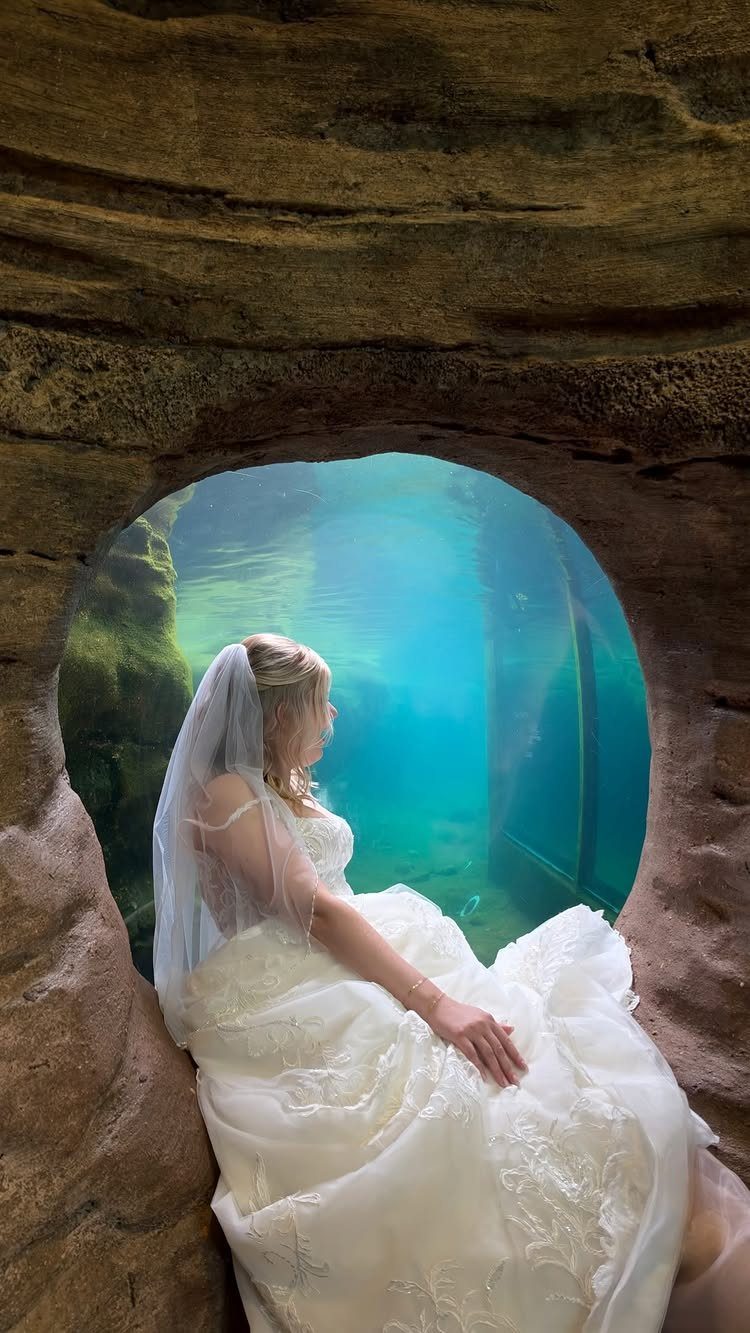 Bride in white dress looking through a circular window into a blue aquarium