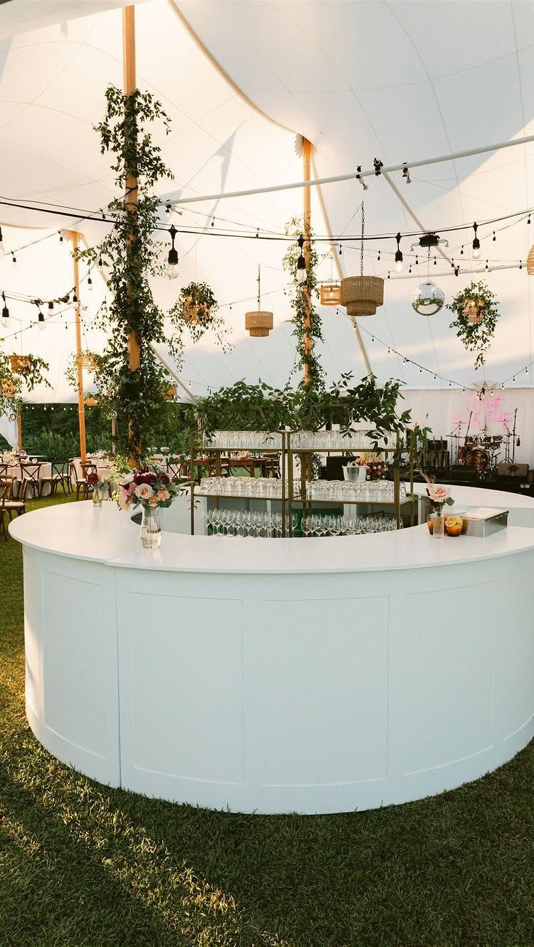 Curved white outdoor bar decorated with flowers, string lights, and hanging greenery.