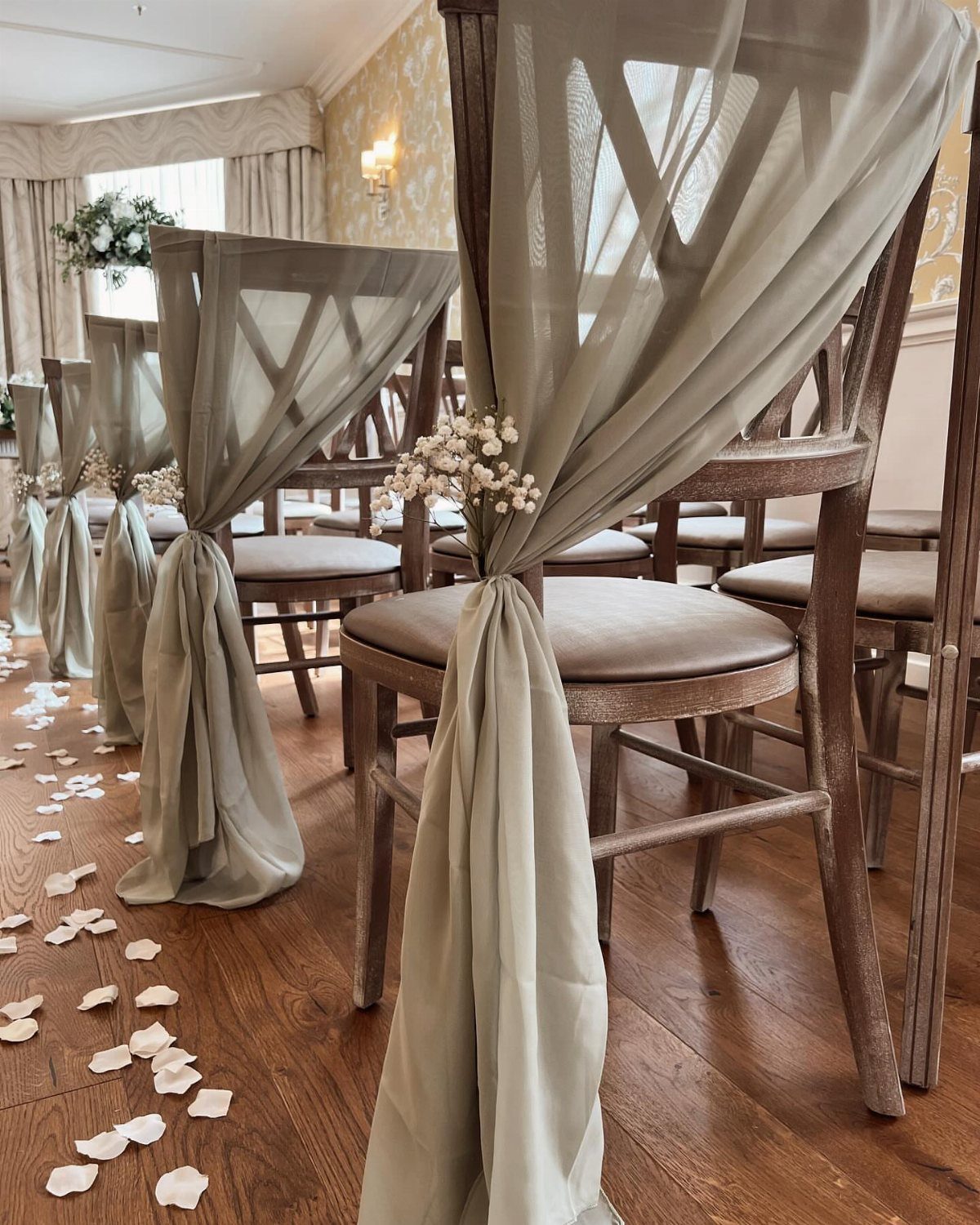 Wooden chairs draped with sage-green fabric and white flowers on a petal-strewn floor.