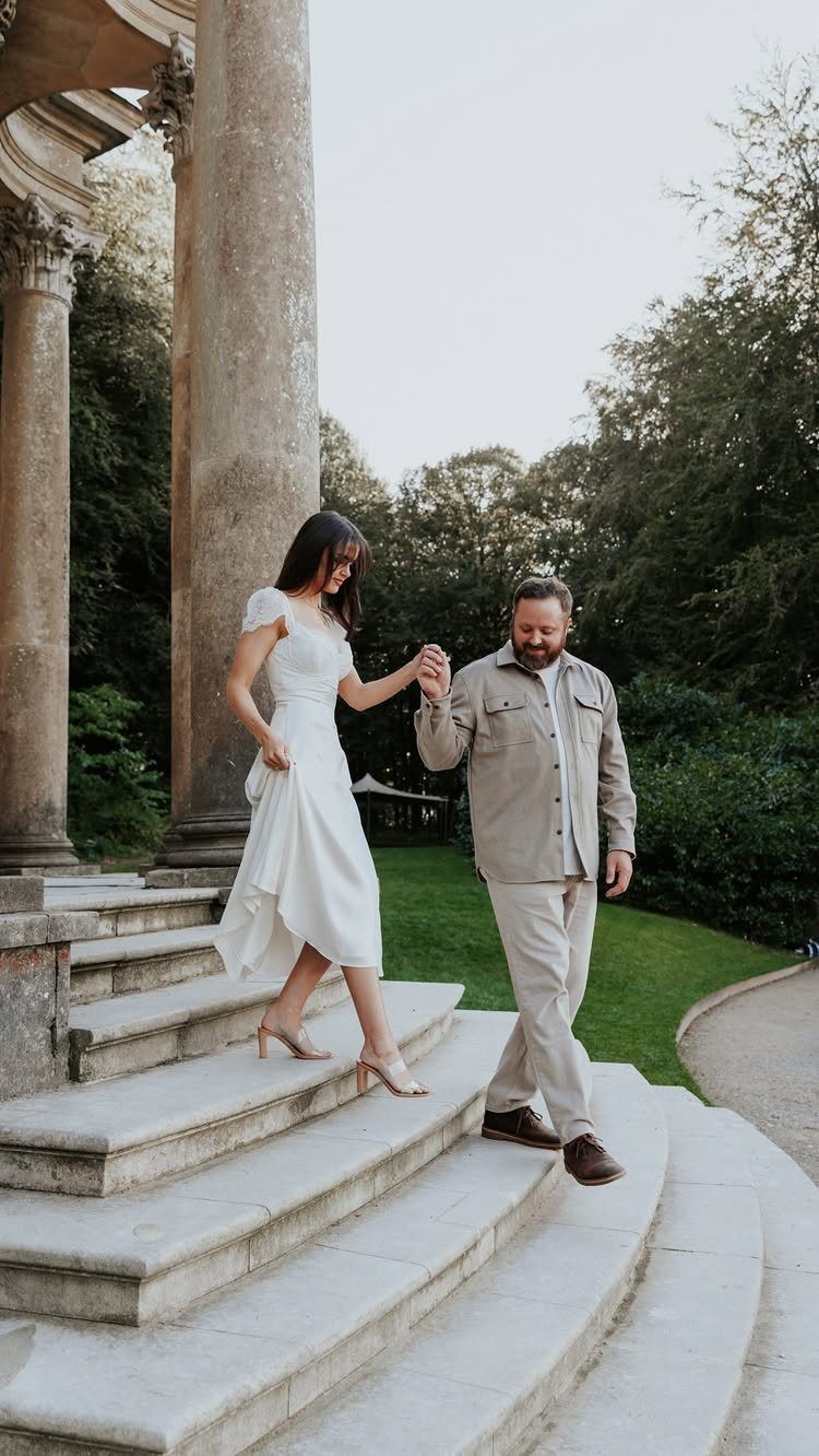 Couple in white and beige attire walking down stone steps of a pavilion.