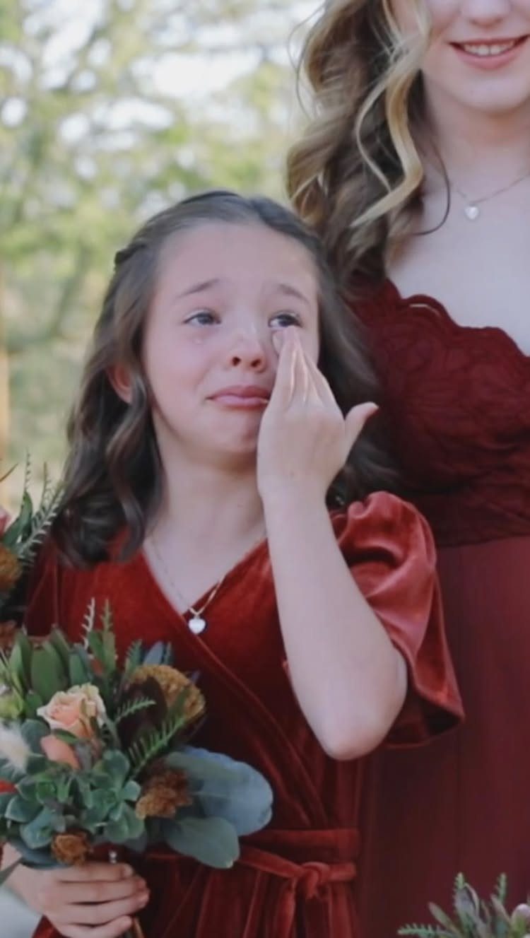 Young girl in red velvet dress wiping a tear while holding flowers.