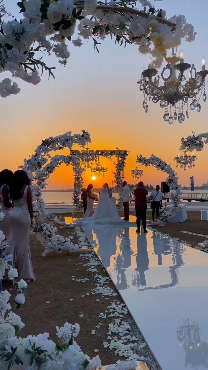 Couple in white wedding attire standing under a floral arch at sunset seaside.