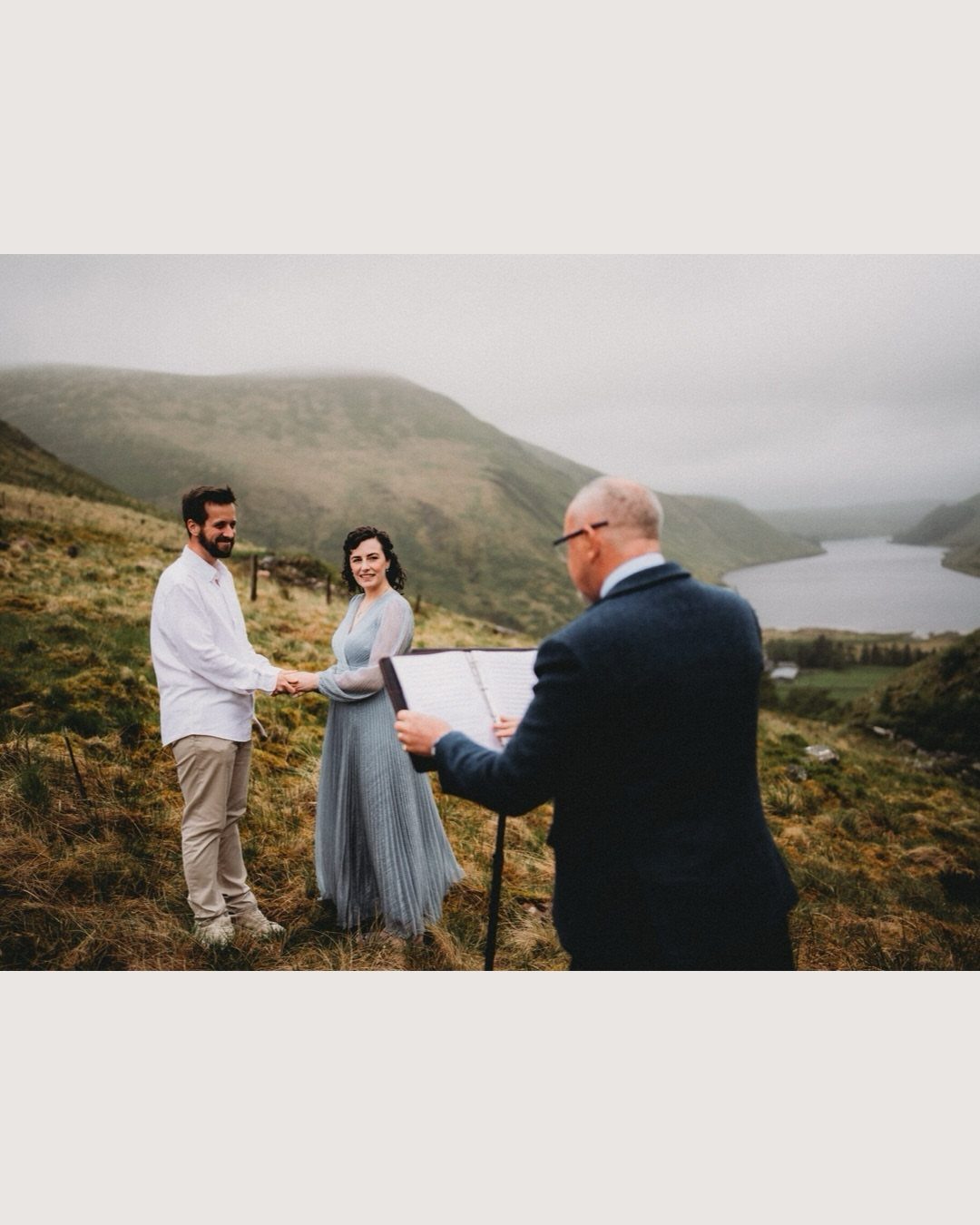 Couple holding hands during a wedding ceremony on a grassy hill by a lake