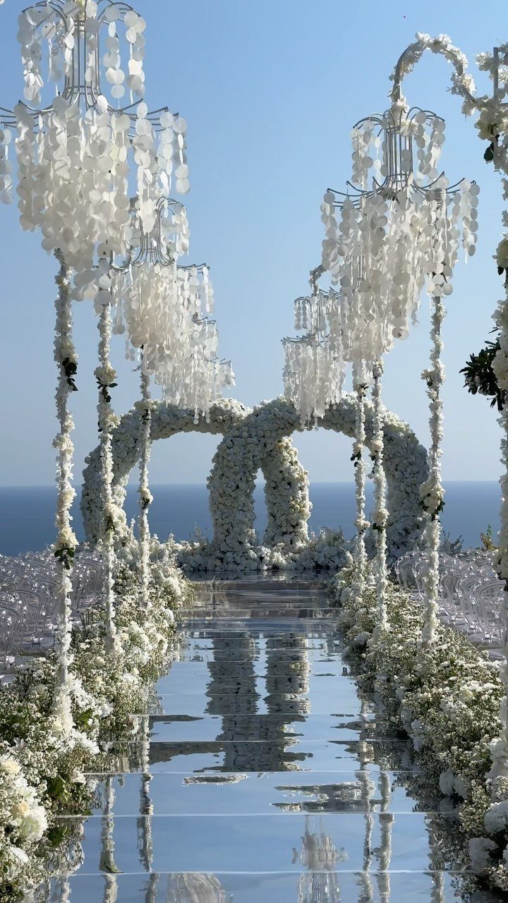 Mirrored aisle with white flowers leading to a floral arch by the ocean.