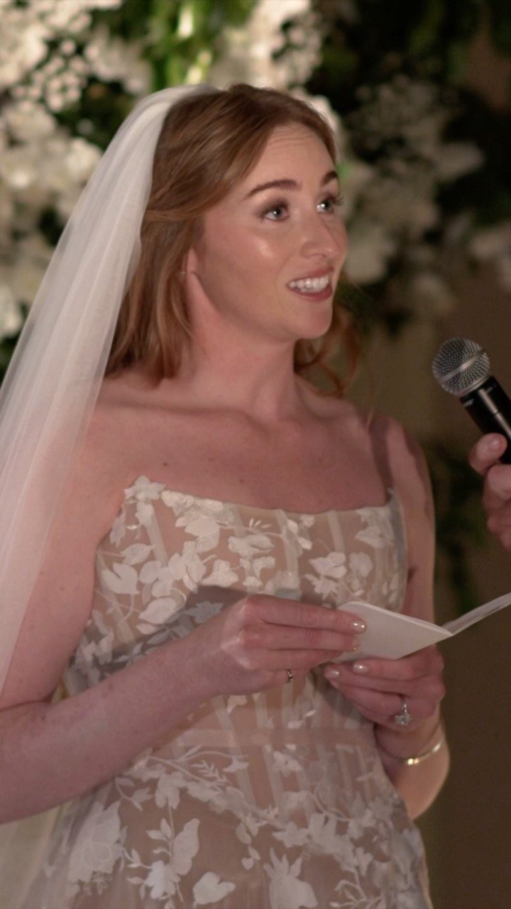 Bride in floral dress and veil reading vows into a microphone