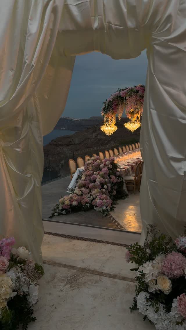 Outdoor dinner table with floral arrangements and chandeliers overlooking a coastal cliffside sunset.